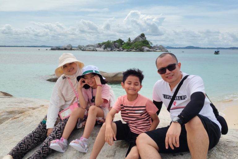 Sebuah Family Yang sedang liburan Paket Tour belitung 2d1n reguler tentang ke pantai laskar pelangi dengan latar belakang langit biru,air jernih yang tenang dan keindahan bebatuan granit .